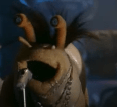 A snail Muppet with piercings and chains sings into a microphone